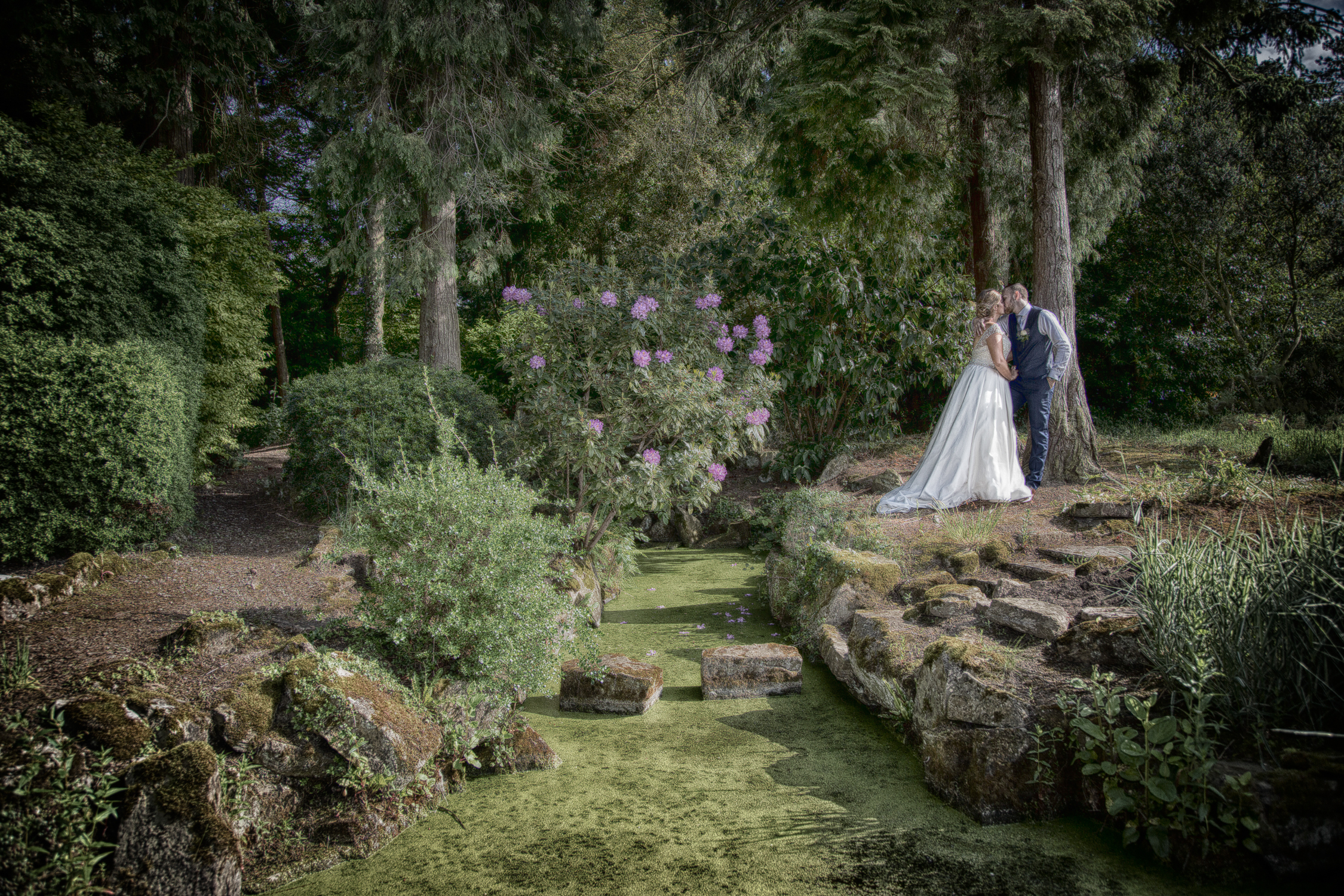 A bride and groom standing next to a pond in a garden.
