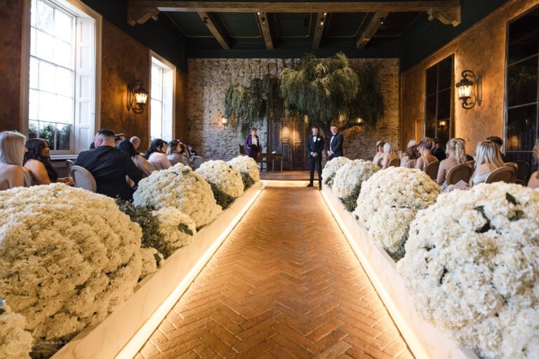 a beautifully decorated indoor wedding ceremony. the aisle is lined with large white floral arrangements, leading to a stone backdrop with two people standing at the altar. guests are seated on either side, in a warmly lit room with tall windows and rustic wooden beams