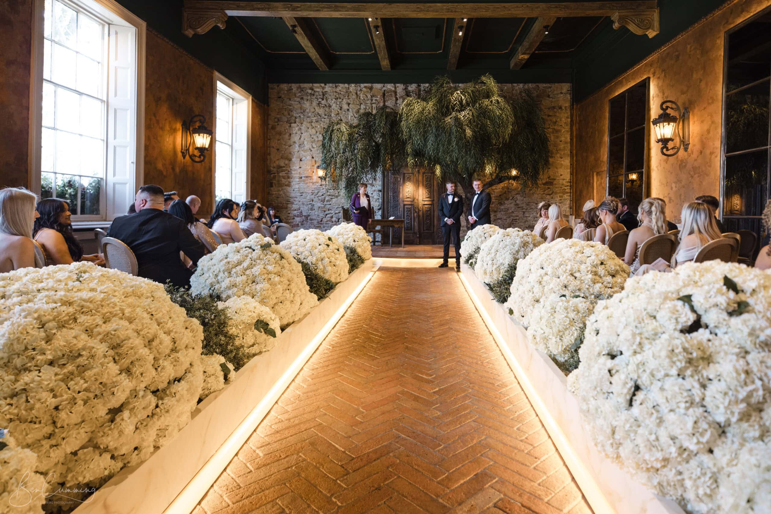 a beautifully decorated indoor wedding ceremony. the aisle is lined with large white floral arrangements, leading to a stone backdrop with two people standing at the altar. guests are seated on either side, in a warmly lit room with tall windows and rustic wooden beams