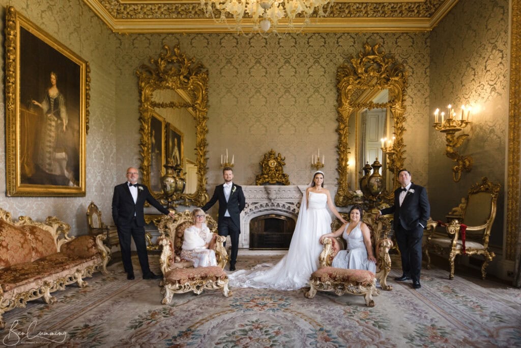 A wedding party poses in an ornate room with gold-framed mirrors, chandeliers, and antique furniture. The bride stands in the center in a white gown, surrounded by the groom and attendants in formal attire. Image by Yorkshire Wedding Photographer - Ben Cumming Photography.