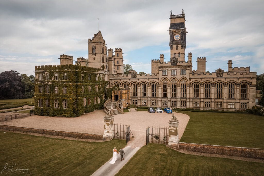 aerial view of a grand historic castle with ivy clad walls, large windows, and an ornate clock tower. a couple in wedding attire walks along the gravel path towards the entrance, surrounded by lush green lawns under a cloudy sky. image by yorkshire wedding photographer ben cumming photography.