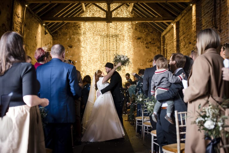 a bride and groom kiss at the altar in a rustic barn wedding venue, surrounded by guests who are standing, some holding flowers and children. warm string lights hang behind the couple, creating a romantic atmosphere. image by yorkshire wedding photographer ben cumming photography.