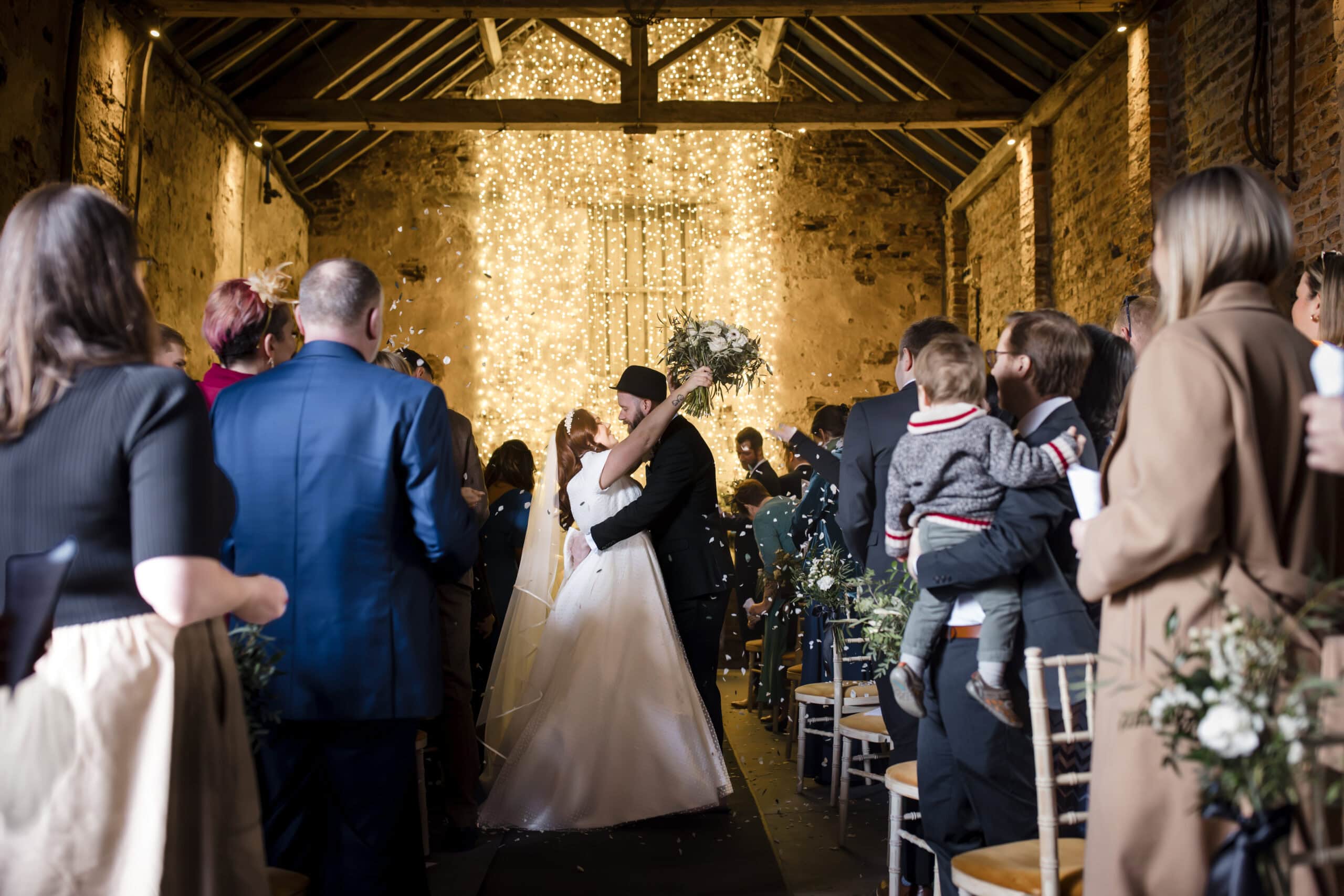 a bride and groom kiss at the altar in a rustic barn wedding venue, surrounded by guests who are standing, some holding flowers and children. warm string lights hang behind the couple, creating a romantic atmosphere. image by yorkshire wedding photographer ben cumming photography.