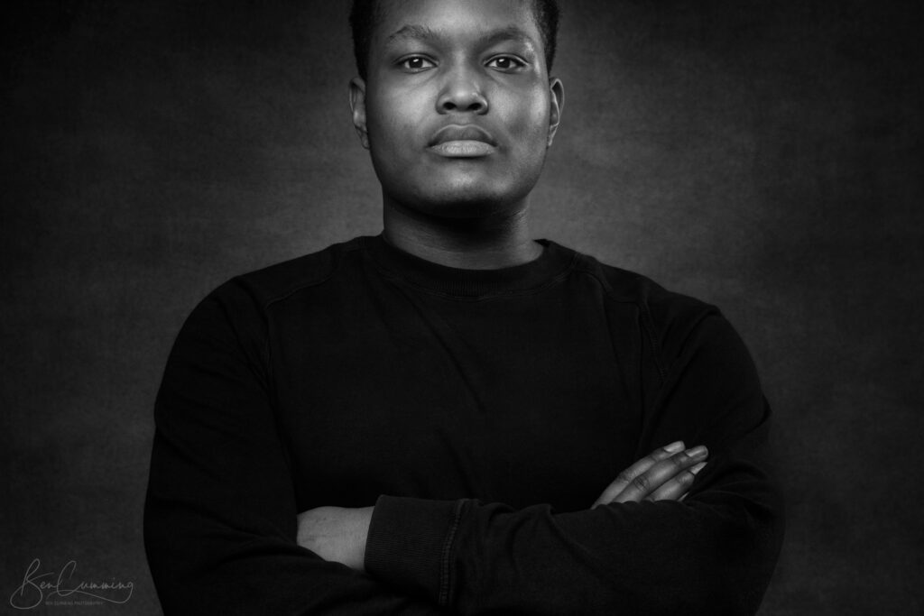 Black and white portrait by a headshot photographer: a person with short hair stands with arms crossed, confidently looking at the camera, in a plain dark sweatshirt against a textured, dark Yorkshire background. Ben Cumming Photography - Wedding Photographer - Yorkshire