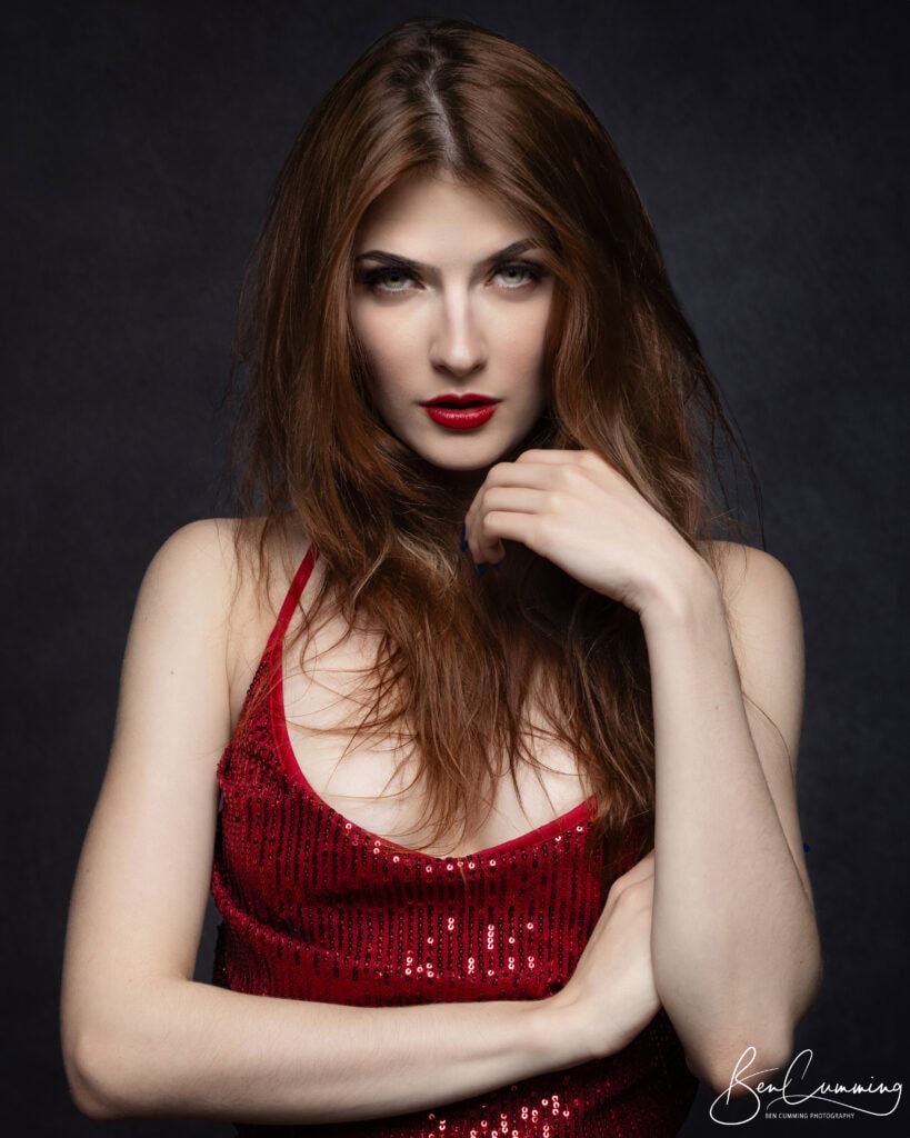 a woman with long brown hair wears a sparkly red sleeveless top. captured by a headshot photographer in yorkshire, she has bold red lipstick, a confident expression, and her hand rests on her hair against a dark background. ben cumming photography wedding photographer yorkshire