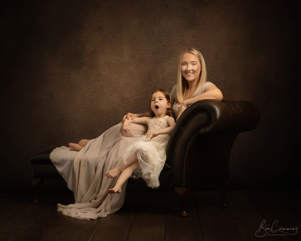 a woman and a young girl sit on a dark chaise lounge against a brown backdrop. captured by a yorkshire family photographer, the woman smiles at the camera while the girl in a white dress looks relaxed, mid yawn or singing. both wear light colored dresses. ben cumming photography wedding photographer yorkshire