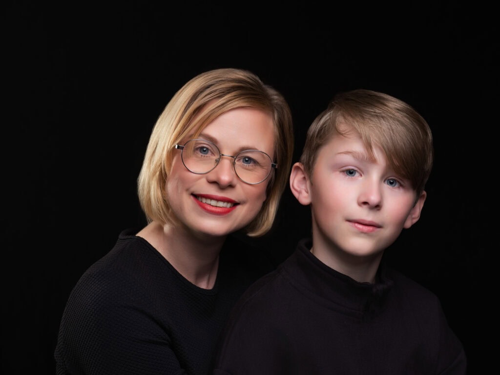 a smiling woman with short blonde hair and glasses sits beside a young boy with light brown hair, both in black tops, against a black background—captured by a yorkshire family photographer. ben cumming photography wedding photographer yorkshire