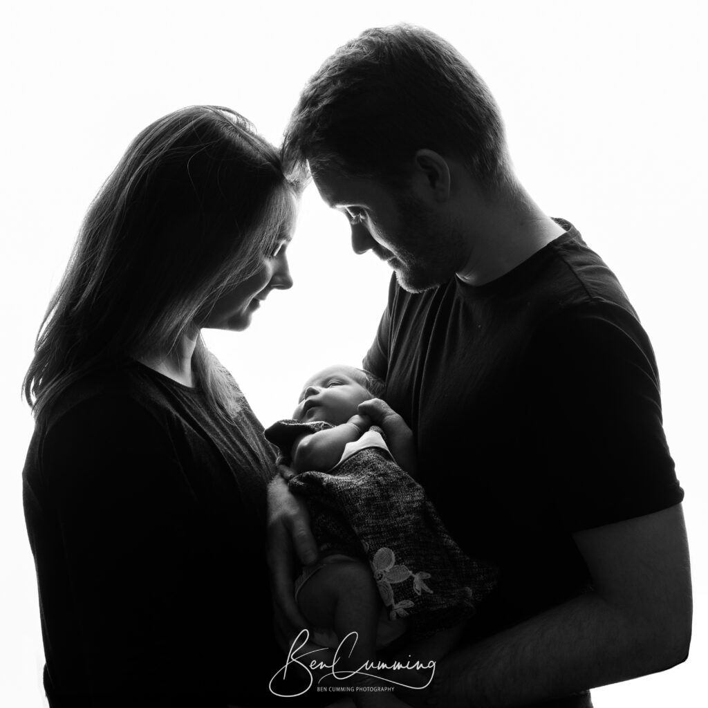 a black and white photo by a yorkshire family photographer shows a couple holding a newborn baby, facing each other and smiling softly, with bright backlighting creating a gentle silhouette effect. ben cumming photography wedding photographer yorkshire
