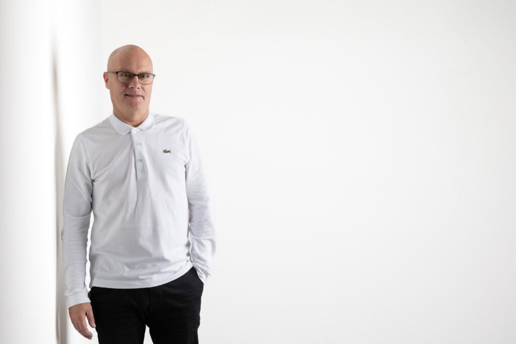 a bald man wearing glasses, a white long sleeve polo shirt, and black pants stands against a plain white background. captured by a yorkshire headshot photographer, he smiles slightly with one hand in his pocket. ben cumming photography wedding photographer yorkshire