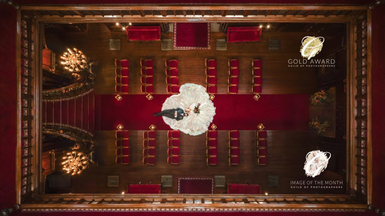 a bride and groom stand in the center of an ornate hall with red carpet and gold chairs, viewed from above. the bride's dress fans out in a circle. this award winning photography is among the finalist images for image of the year