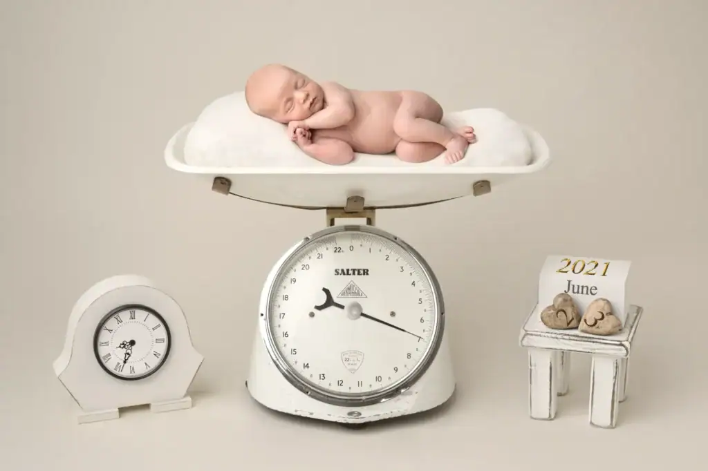 a smiling newborn baby lies on a pillow atop a vintage set of scales. next to the scales are a small clock and a calendar displaying "2021 june" with two wooden heart decorations. the background is plain and light coloured. ben cumming photography wedding photographer yorkshire