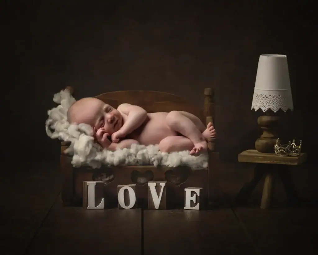 a newborn baby sleeps curled up on a small wooden bed with a fluffy white blanket. in front of the bed, wooden blocks spell “love.” a tiny lamp and a small crown sit on a wooden stool nearby. ben cumming photography wedding photographer yorkshire