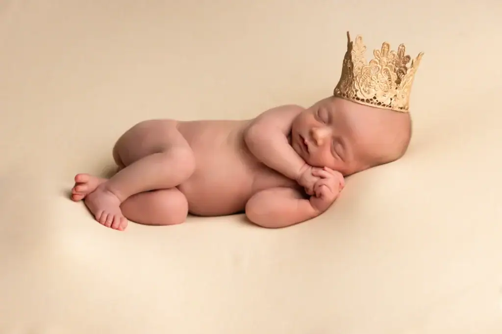 a newborn baby sleeps peacefully on a light beige surface, wearing a delicate gold crown on their head, with hands tucked under their cheek and legs curled up. ben cumming photography wedding photographer yorkshire