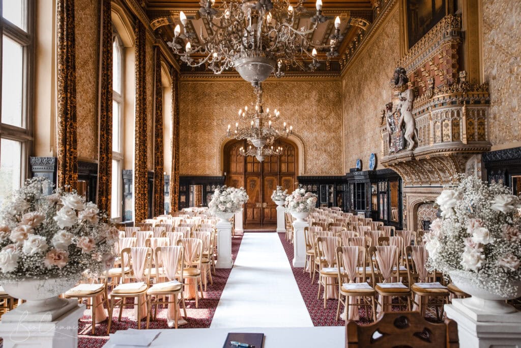a lavishly decorated ballroom set up for a wedding ceremony at carlton towers, featuring rows of wooden chairs, large white floral arrangements, ornate chandeliers, gold walls, tall windows, and an elaborate fireplace—perfect for a wedding photographer. ben cumming photography wedding photographer yorkshire