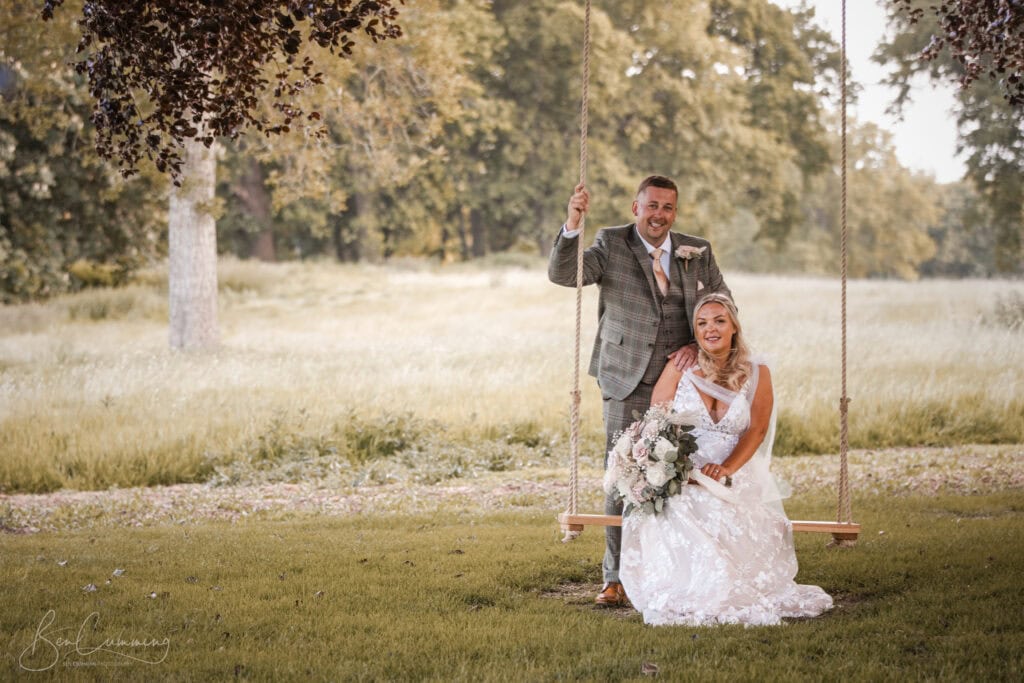 a bride in a white lace dress sits on a wooden swing holding a bouquet, while her groom in a grey checked suit stands behind her, both smiling—captured beautifully by a carlton towers wedding photographer in a grassy outdoor setting with trees. ben cumming photography wedding photographer yorkshire