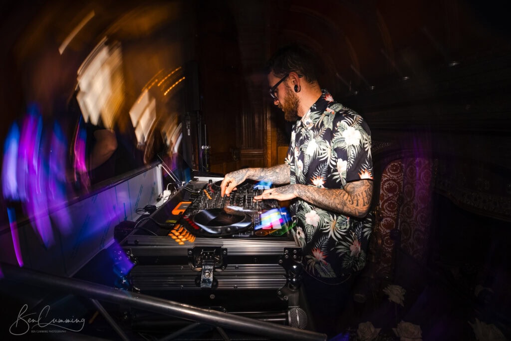 a bearded man with glasses and tattoos, wearing a floral shirt, is djing at a console with colourful lights in a dimly lit venue—a scene any carlton towers wedding photographer would love, with motion blur adding to the dynamic atmosphere. ben cumming photography wedding photographer yorkshire