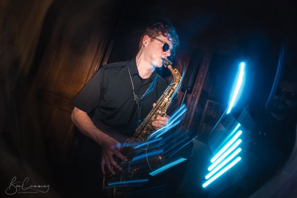 a musician wearing sunglasses plays a saxophone passionately, surrounded by dynamic blue light trails, creating an energetic and lively atmosphere—captured with the artistic eye of a carlton towers wedding photographer—in a dimly lit room. ben cumming photography wedding photographer yorkshire
