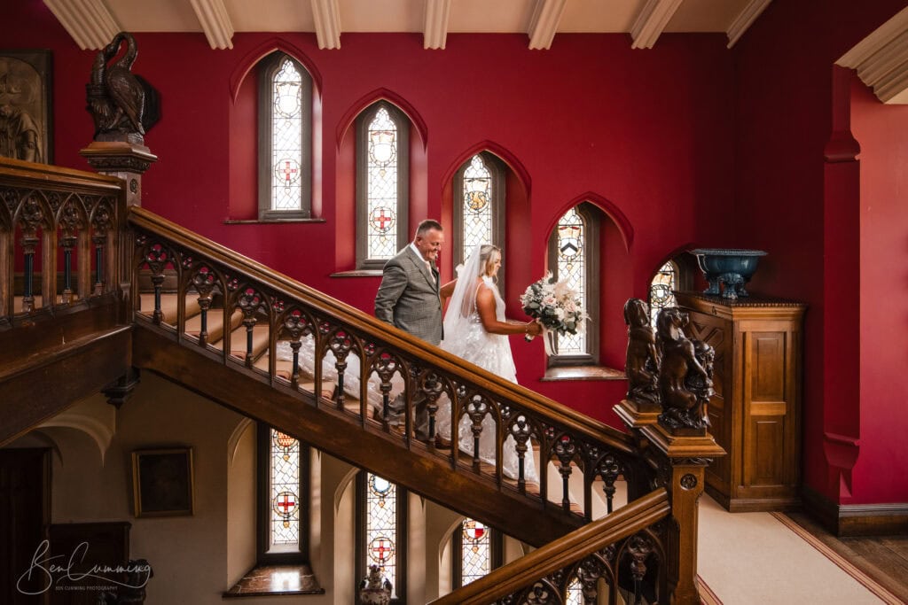 a bride and groom stand on an ornate wooden staircase inside a historic building with red walls and stained glass windows, smiling at one another. captured by a carlton towers wedding photographer, the moment radiates elegance and joy. ben cumming photography wedding photographer yorkshire
