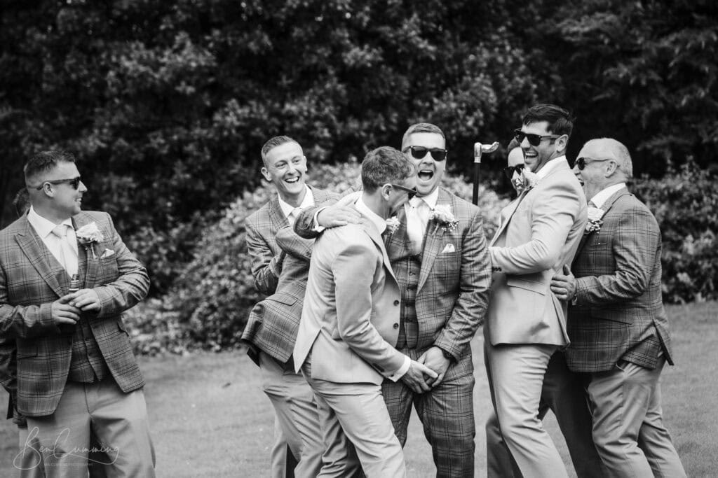 a group of men in suits, laughing and embracing each other outdoors. captured by a carlton towers wedding photographer, they appear to be at a formal event, possibly a wedding, standing on grass with trees in the background. ben cumming photography wedding photographer yorkshire