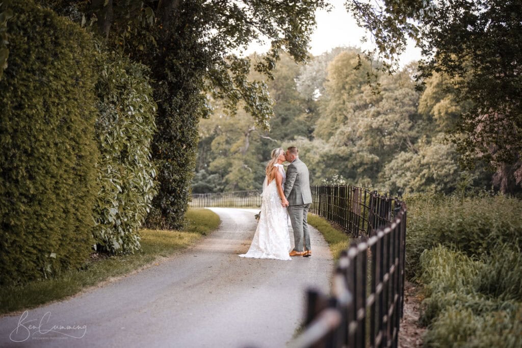 a bride and groom stand on a tree lined path, facing each other and holding hands. captured by a carlton towers wedding photographer, they’re surrounded by greenery, a black iron fence, and a lush wooded background. ben cumming photography wedding photographer yorkshire