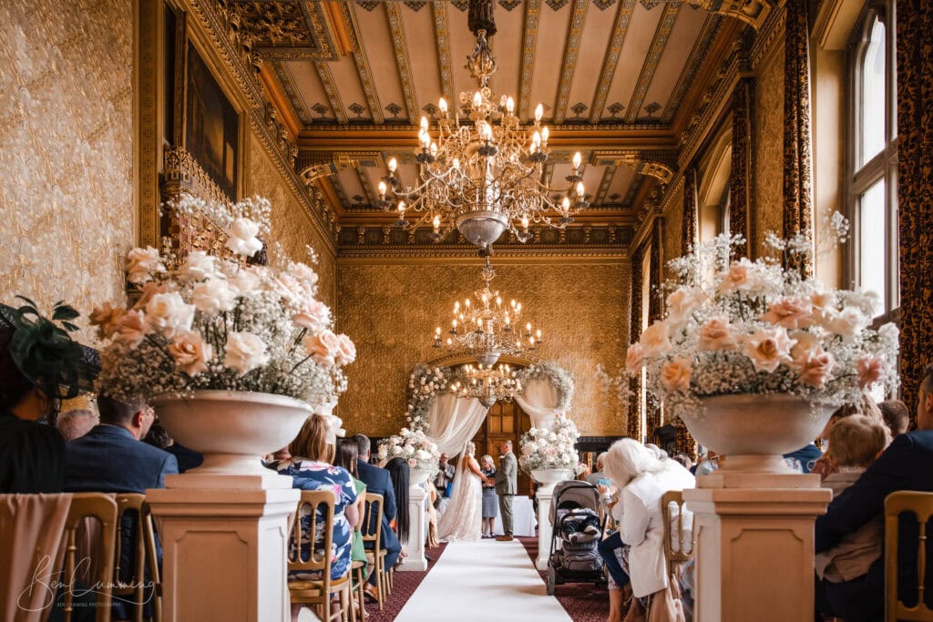 a carlton towers wedding photographer captures a beautiful ceremony in an ornate, gold decorated hall with chandeliers, floral arrangements lining the aisle, and guests seated on either side watching the couple at the altar. ben cumming photography wedding photographer yorkshire