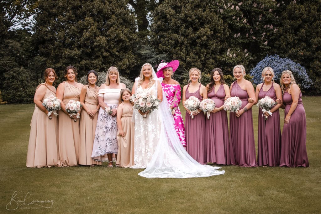 a bride in a white wedding dress stands outdoors with nine women and a young girl, all holding bouquets. captured by a carlton towers wedding photographer, the group poses among trees and flowers, most in mauve or beige dresses. ben cumming photography wedding photographer yorkshire