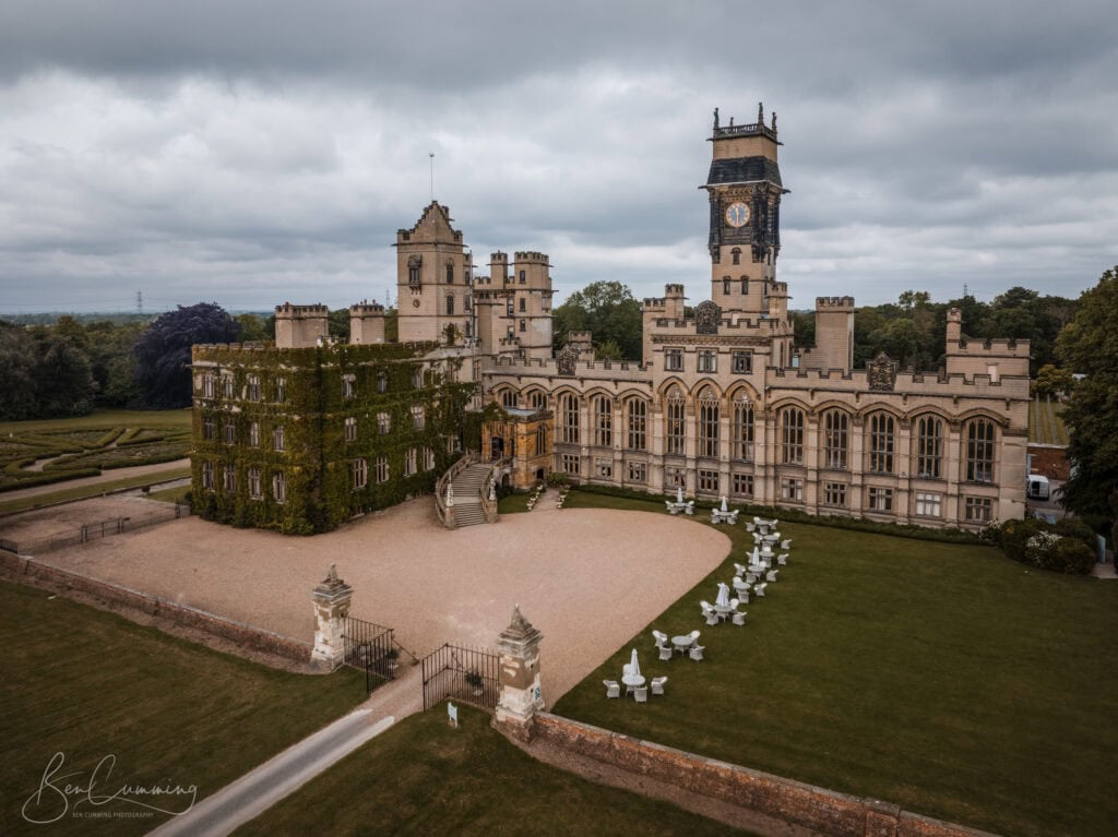 aerial view of a large, historic castle with a clock tower, surrounded by green lawns and garden chairs. the building features ornate architecture and ivy covered walls under a cloudy sky. ben cumming photography wedding photographer yorkshire