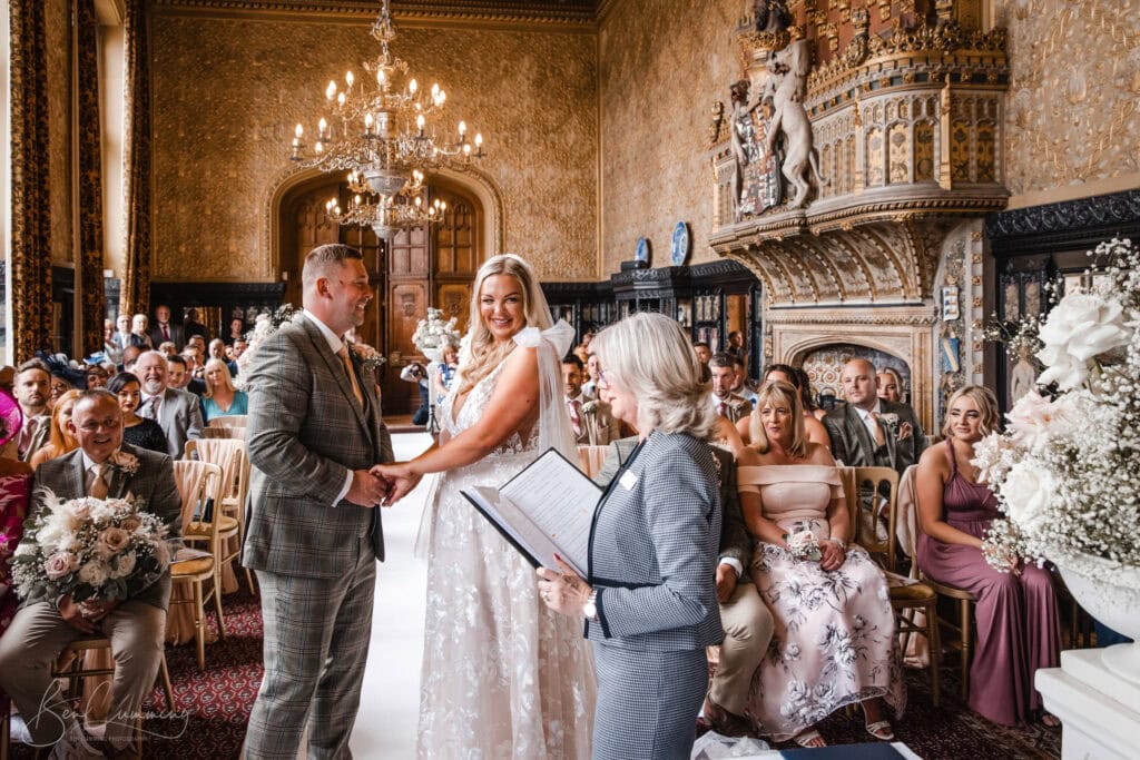 a bride and groom stand holding hands, smiling during their ceremony in the ornate carlton towers, with chandeliers, floral arrangements, and guests seated, as their wedding photographer captures the moment while an officiant reads from a book. ben cumming photography wedding photographer yorkshire