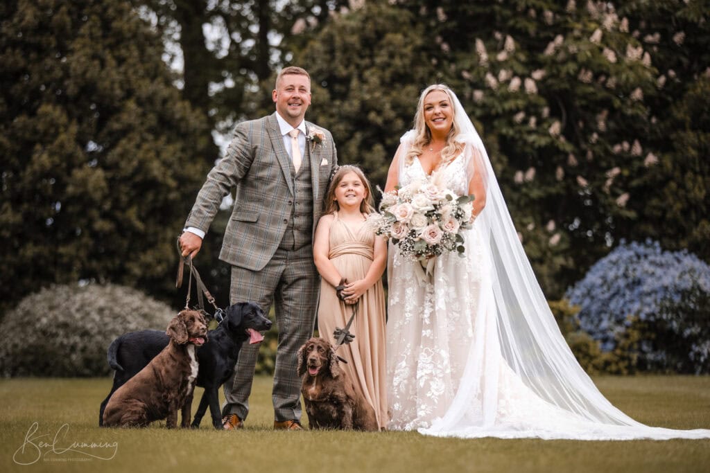 a bride in a white gown, groom in a checked suit, and young girl in beige stand smiling on grass with three dogs on leads, surrounded by trees—captured beautifully by a carlton towers wedding photographer. ben cumming photography wedding photographer yorkshire