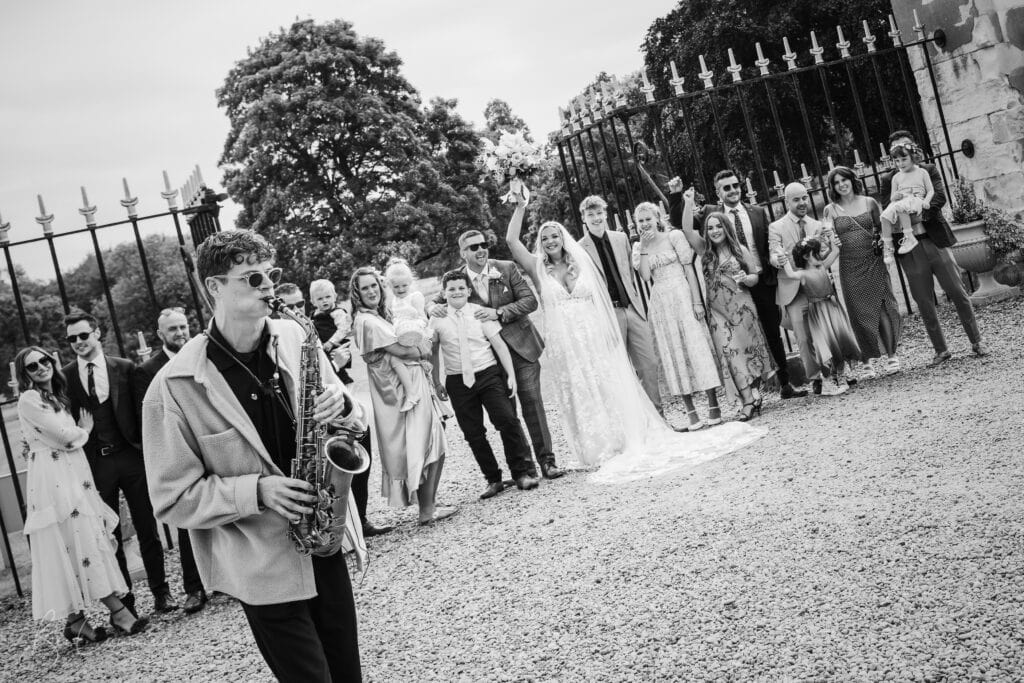 a black and white photo by a carlton towers wedding photographer captures a lively wedding party outdoors. a saxophonist plays in the foreground as the bride, groom, and guests pose and cheer near a wrought iron fence and trees. ben cumming photography wedding photographer yorkshire