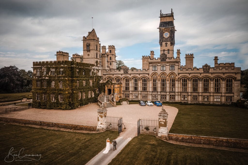 a grand, historic castle with ivy covered walls and a central clock tower stands amid manicured lawns. a carlton towers wedding photographer captures the bride and groom as they walk through the entrance gate, several cars parked outside. ben cumming photography wedding photographer yorkshire