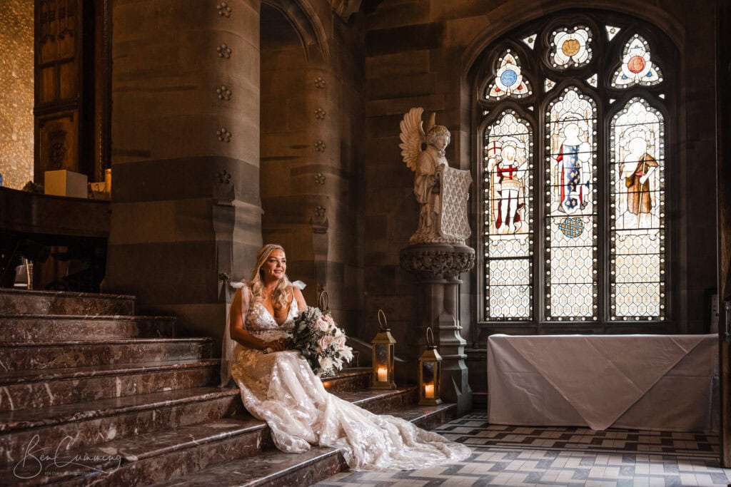 a bride in a white lace dress sits on marble steps in a grand, stone walled room with stained glass windows, holding a bouquet and smiling softly. candles and an angel statue are nearby—a perfect scene for a carlton towers wedding photographer. ben cumming photography wedding photographer yorkshire