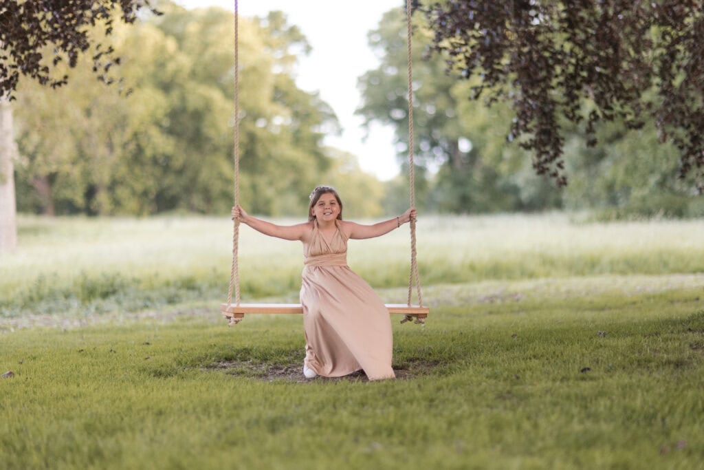 a girl in a long, light pink dress is sitting and smiling on a wooden swing hanging from tree branches in a green, grassy field—an enchanting scene perfect for any carlton towers wedding photographer. ben cumming photography wedding photographer yorkshire