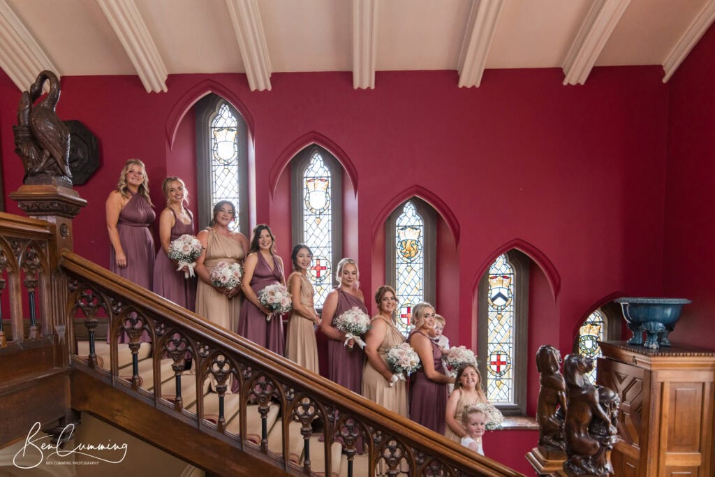 eleven bridesmaids in mismatched blush and mauve dresses stand on a wooden staircase at carlton towers, framed by a red wall and stained glass windows. each holds white flowers, while a young girl peeks out from the bottom right—perfect for any wedding photographer. ben cumming photography wedding photographer yorkshire