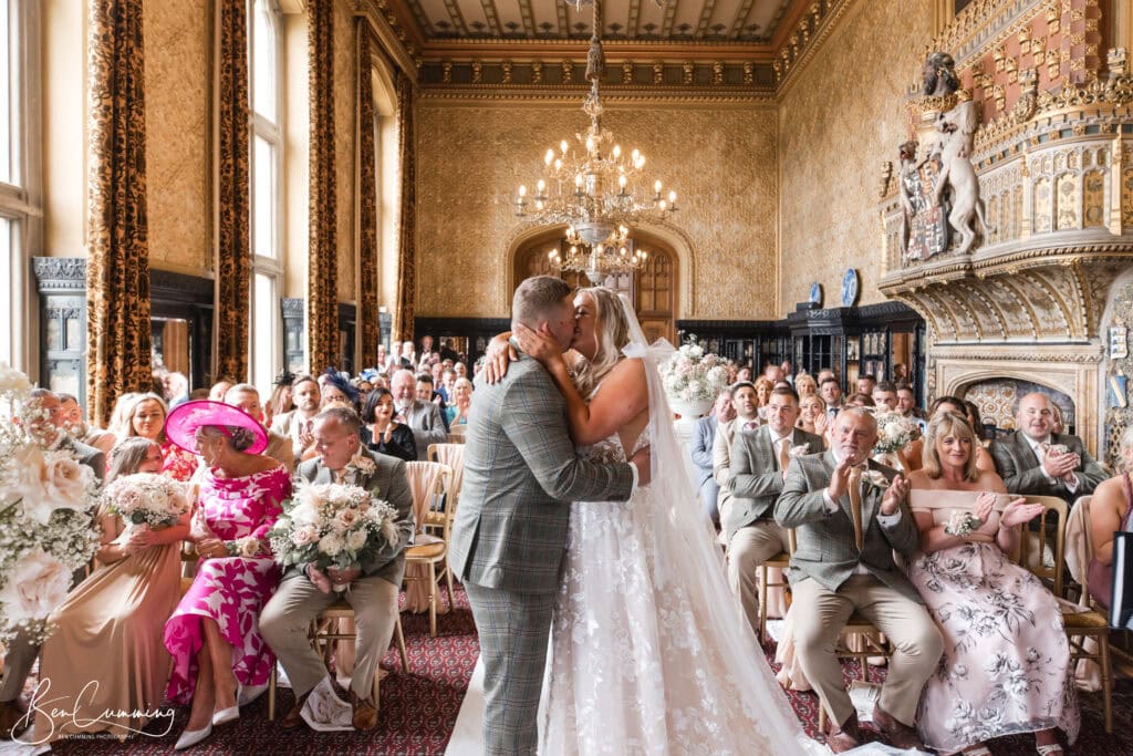 a bride and groom kiss at the altar during their wedding ceremony in an ornate, gold decorated room, whilst guests on both sides celebrate—beautifully captured by a carlton towers wedding photographer. ben cumming photography wedding photographer yorkshire