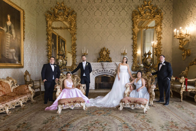 six people in formal attire pose in a lavish, ornate room with gilt framed mirrors, a fireplace, and antique furniture—a scene perfectly captured by an experienced allerton castle wedding photographer. two women wear dresses, one white and one pink, while four people wear dinner jackets.