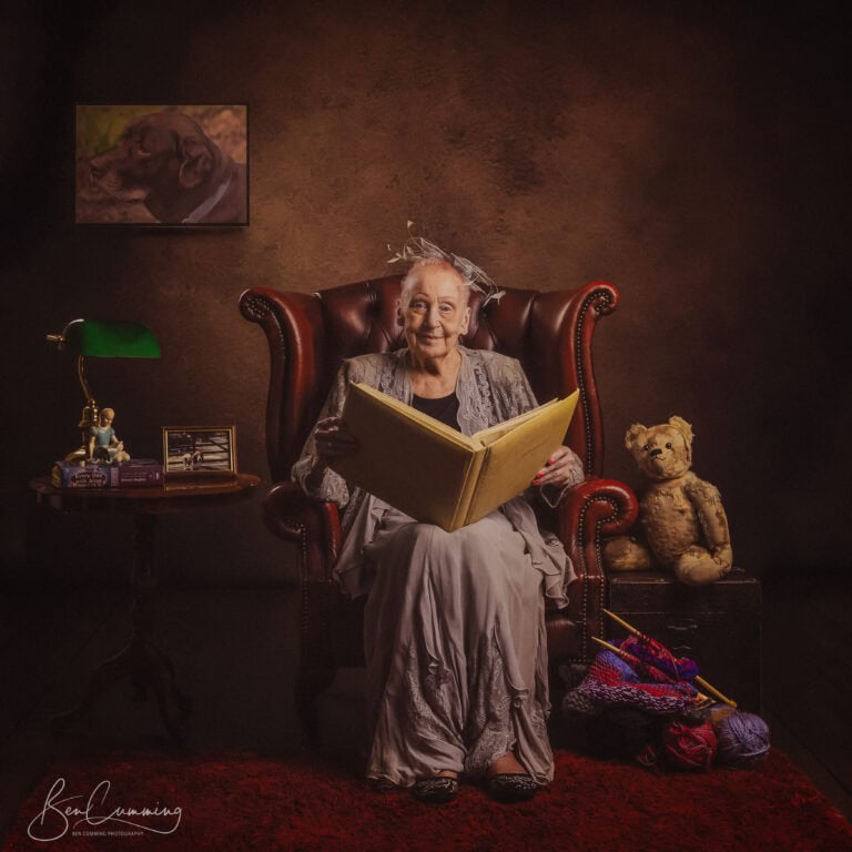 Ben Cumming's mum sits in a leather armchair, smiling and holding a large yellow book—making memories that will never be forgotten. a teddy bear and knitting supplies are by her side, while a framed dog photo hangs on the wall behind her.
